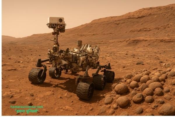 NASA का Curiosity रोवर (क्यूरियोसिटी रोवर) मंगल ग्रह की सतह पर खड़ा है, पास में गोल पत्थर जैसी संरचनाएँ दिखाई दे रही हैं।